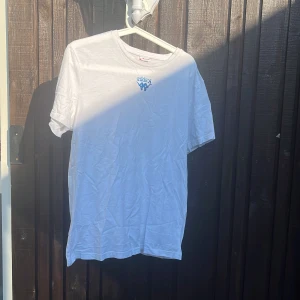 Vit Kappa t-shirt med logga - Vit t-shirt från Kappa med blå logga på bröstet och grå axelband med svarta Kappa-symboler. Klassisk rund hals och korta ärmar. Perfekt för en sportig och avslappnad stil.