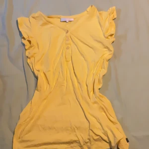 Gul blus med volangärm från Zeze - Snygg gul blus från Zeze Nordic i mjuk bomull. Modellen har korta volangärmar och knappar framtill som ger en söt touch. Perfekt för dig som gillar färg och vill sticka ut lite extra. Superfin till jeans eller kjol!