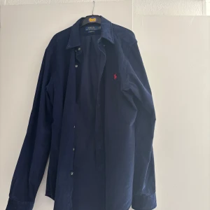 Mörkblå manchesterskjorta Polo Ralph Lauren - Snygg mörkblå manchesterskjorta från Polo Ralph Lauren med klassisk krage och knappar framtill. Skjortan har lång ärm och den ikoniska röda loggan broderad på bröstet.  Passar M och L: slimfit och är väldigt bra skick nytt 
