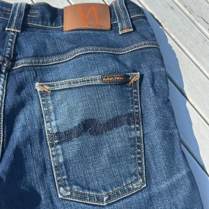 Blå jeans från Nudie Jeans - Snygga blå jeans från Nudie Jeans med klassisk femficksdesign och kontrastsömmar. Jeansen har en rak passform och är tillverkade i slitstarkt bomullsjeans. Bakfickorna har Nudie Jeans ikoniska broderi och läderpatch i midjan. Med en stilren slitning på benet! Strl: W32, L32.