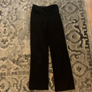 Svarta kostymbyxor med raka ben - Snygga svarta kostymbyxor med raka, lite vidare ben och klassiska hällor för bälte. Byxorna har en stilren look med diskret dragkedja och knapp framtill. Perfekta för en clean och dressad outfit.