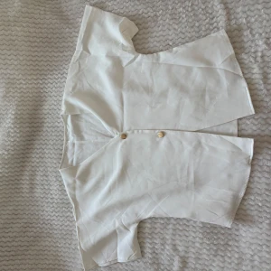 Vit blus med knappar - Stilren vit blus med korta ärmar och två dekorativa guldfärgade knappar framtill. Blusen har en rak passform och är tillverkad i ett lätt och mjukt tyg som ger en luftig känsla. Perfekt för en fräsch och enkel look.