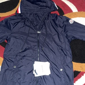 Marinblå vindjacka från Moncler - Snygg marinblå vindjacka från Moncler med huva och dragkedja framtill. Jackan har Moncler-logga på ärmen, snörning i huvan och praktiska fickor med knappar. Tillverkad i lätt och glansig polyester, perfekt för blåsiga dagar.