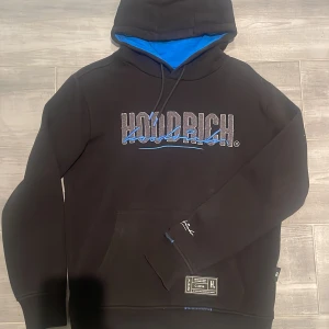 Svart hoodie från Hoodrich med blå detaljer - Svart hoodie från Hoodrich med broderad logga i grått och blått framtill och baktill. Insidan av huvan är blå, och tröjan har känguruficka och snörning i huvan. Snygga detaljer och tryck på ärm och nederkant. Aldrig använd!