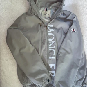 Grå vindjacka från Moncler med huva - Snygg grå vindjacka från Moncler med huva och dragkedja framtill. Stor logga framtill intill dragkedjan och klassisk moclere märke på ärmen. Jackan har huva, dragkedja, bröstficka med zip och elastiska muddar. Perfekt för dig som vill ha premiumkänsla på sena sommarkvällar och både höst- och vårdagar. 