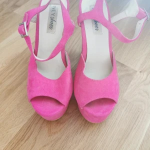 Rosa mockasandaler med klack från NLY Shoes - Snygga sandaler i stark rosa mocka med hög klack och plattform. Skorna har öppen tå, bred rem över foten och justerbart spänne vid ankeln. Perfekta för dig som vill sticka ut med färg och höjd. Klackhöjd ca 11 cm.