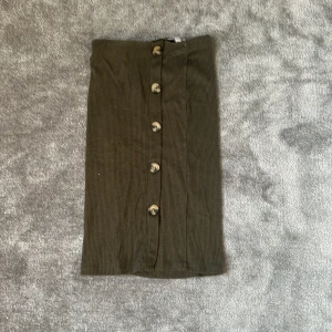 Olivgrön ribbad kjol med knappar - Olivgrön ribbad kjol från Skill med snygga knappar framtill. Kjolen har en rak passform och är tillverkad i ett mjukt, stretchigt material som sitter skönt. Perfekt för dig som gillar stilrena och enkla plagg med en twist.