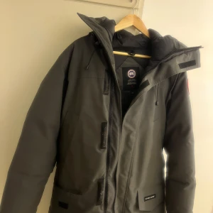 Mörkgrå dunjacka från Canada Goose - Säljer en mörkgrå dunjacka från Canada Goose med huva och dragkedja. Jackan har en clean look med diskreta fickor och logga på ärmen. Insidan är quiltad för extra värme och komfort. Perfekt för kalla dagar när du vill hålla stilen. Man kan även ha päls i luvan det finns en zip som gömmer sig. Passar även i folk med storlek L.