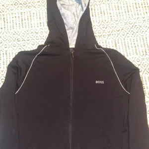 Svart hoodiejacka från BOSS - Svart hoodiejacka från BOSS med dragkedja framtill och vit piping längs axlarna. Jackan har huva med grått innerfoder och diskret BOSS-logga på bröstet. Tillverkad i mjuk bomull, perfekt för en chill och stilren look. Tunn hoodie, Skick 10/10 använd fåtal gånger lite solbränd i vissa ljus 