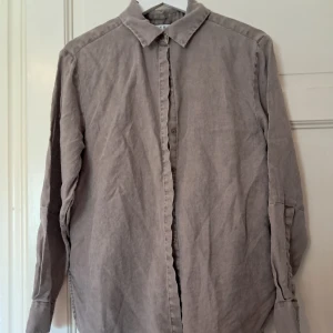 Stenfärgad linneskjorta från Wera - Stilren och oversized skjorta från Wera i en mjuk grå nyans. Skjortan är tillverkad i linne, har klassisk krage, knäppning framtill och långa ärmar med manschett. Perfekt för en avslappnad och trendig look.