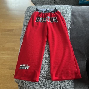 Röda sportiga byxor från SHEIN - Röda sportiga byxor från SHEIN med bred resår i midjan där det står SPORT. Byxorna har tryck med texten BOSTON framtill och USA Energetic på benet. De är vida i modellen och tillverkade i ett mesh-liknande syntetmaterial.
