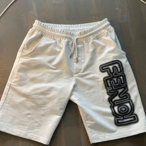 Vita Fendi shorts med logga - Snygga vita shorts från Fendi i bomull med elastisk midja och snörning. Stora Fendi-loggan i svart och grått tryck längs ena benet ger en cool streetkänsla. Perfekta för sommaren och chill dagar. Två sidofickor och relaxed fit.