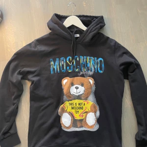Svart Moschino hoodie med björntryck - Cool svart hoodie från Moschino med stort färgglatt tryck av en nallebjörn och texten 'This is not a Moschino toy' på magen. Oversized känsla, huva med snörning och tillverkad i 100% bomull. Perfekt statement-plagg för dig som gillar streetstyle.