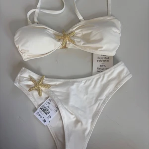 Vit bikini med sjöstjärnedetaljer - Superfin vit bikini med axelband och glittriga guldfärgade sjöstjärnor både på överdelen och trosan. Tillverkad i återvunnen polyester som känns mjuk mot huden. Perfekt för strandhäng och poolparty i sommar!