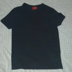 Svart t-shirt från Hugo, klassisk stil - Svart t-shirt från Hugo med enkel och clean design. T-shirten har rund halsringning, korta ärmar och diskret Hugo-logga i nacken. Tillverkad i mjuk bomull som känns skön mot huden. Perfekt för dig som gillar stilrena basplagg.