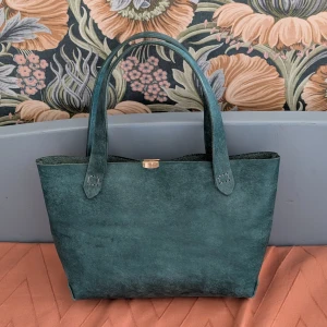 Turkos skinnväska med guldspänne - Snygg turkos handväska i skinn med stilren, fyrkantig form och två rejäla handtag. Väskan har ett diskret guldspänne upptill och är perfekt för dig som gillar minimalistisk design och vill ha plats för allt du behöver under dagen. OBS! Unik handgjord väska