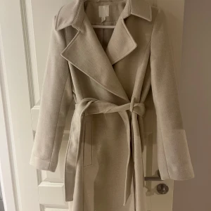 Beige kappa med bälte från H&M - Stilren beige kappa från H&M med bred krage och knytskärp i midjan. Lång modell med raka ärmar, dolda knappar och två fickor framtill. .