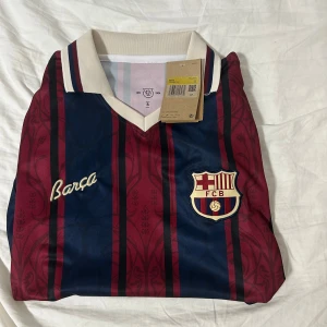 FC Barcelona Ronaldinho 10 tröja - Retroinspirerad FC Barcelona fotbollströja med Ronaldinho och nummer 10 på ryggen. Tröjan har klassiska blå och vinröda ränder, gul text, broderad Barça-logga och klubbmärke. Vit krage med rödblå detaljer. Tillverkad i lätt och ventilerande material. Priset går att diskutera storlek S