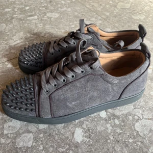 Gråa Louboutin sneakers med nitar - Snygga grå sneakers från Christian Louboutin i mocka med ikoniska röda sulor. Skorna har rund tå, snörning och coola svarta nitar på tån för en edgy vibe. dem är också köpta på plick !! säljer min storebrors skor eftersom att dem aldrig kommit till användning pga för liten storlek 🩶