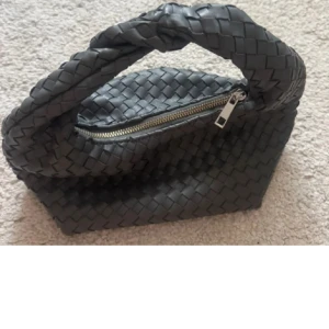 Gina flätad väska  - Säljer en handväska med snygg flätad design och twistat handtag. Väskan är gjord i skinn och har dragkedja upptill. Perfekt storlek för det viktigaste och ger en cool vibe till din outfit.