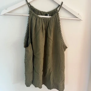 Olivgrön linnetopp  - Snygg olivgrön linnetopp med tunna axelband och dekorativa spetsbollar längs ärmhålen. Toppen har en avslappnad passform och knäppning bak i nacken. Perfekt för varma dagar och enkel att styla med jeans eller kjol.