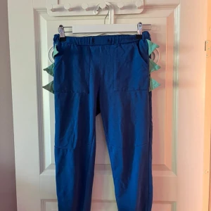 Blå mjukisbyxor med drakdetaljer BARN 140 - Blå mjukisbyxor med elastisk midja och stora fickor framtill. Sidorna har gröna taggiga detaljer som ger en cool draklook. Perfekta för dig som vill sticka ut och gillar unika plagg.