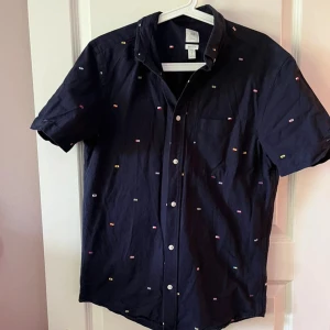 Mörkblå kortärmad skjorta med mönster - Snygg mörkblå skjorta med korta ärmar och små färgglada broderade detaljer över hela tyget. Skjortan har klassisk krage, knäppning framtill och en bröstficka. Perfekt för dig som vill ha en stilren men ändå lekfull look.