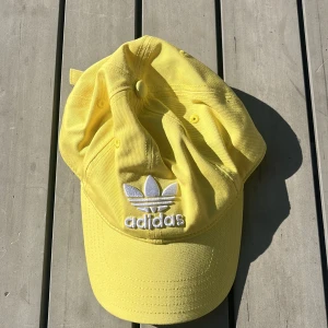 Gul keps från Adidas med broderad logga - Snygg gul keps från Adidas med klassisk vit broderad trefoil-logga framtill. Justerbar rem baktill för perfekt passform. Tillverkad i bomull, perfekt för soliga dagar och streetstyle. Enkel och stilren design som lyfter vilken outfit som helst.