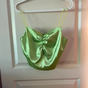 Grön glansig linnetopp med rynk - Snygg grön linnetopp i glansigt material med tunna axelband och rynkdetalj framtill. Perfekt för dig som vill sticka ut med en färgstark och trendig look. Toppen har en croppad passform och är superlätt att matcha med jeans eller kjol.