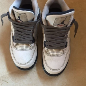 Nike Air Jordan 4 Retro vit/grå - Säljer ett par Nike Air Jordan 4 Retro sneakers i vitt skinn med grå och svarta detaljer samt röda accenter på sulan. Klassisk meshpanel på sidorna, tjock plös med Jumpman-logga och breda grå skosnören. Perfekta för dig som gillar ikoniska basketskor.