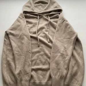 Beige stickad hoodie med huva - Snygg beige stickad hoodie med huva och snörning. Tröjan har långa ärmar och ribbade muddar vid ärmslut och nederkant. Perfekt för en chill och avslappnad stil.