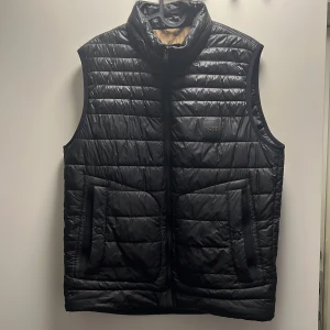 Svart dunväst från BOSS, stl 48 - Stilren svart dunväst från BOSS med quiltad design och hög krage. Västen har två fickor med dragkedja framtill och diskret BOSS-logga på bröstet. Insidan är fodrad i beige och orange detaljer. Perfekt för lager-på-lager och snygg till hoodie eller tröja.