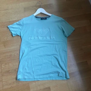 Ljusblå Emporio Armani t-shirt - Snygg ljusblå t-shirt från Emporio Armani med klassisk logga och text i relief på bröstet. T-shirten har rund halsringning och korta ärmar. Tillverkad i mjuk bomull som känns skön mot huden. Perfekt för en clean och stilren look.