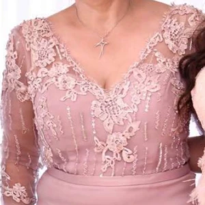Ljusrosa långklänning med spetsdetaljer - Elegant ljusrosa långklänning med v-ringning och trekvartsärm. Överdelen har vackra spetsapplikationer och blommiga broderier, samt transparenta ärmar. Kjolen är slät och figurnära. Perfekt för speciella tillfällen.