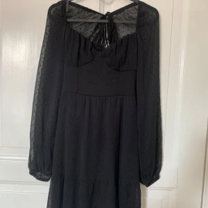 Svart långklänning med prickiga ärmar - Supersnygg svart långklänning med transparenta, långärmade ärmar i prickigt tyg. Klänningen har markerad byst, knytning i ryggen och flowy kjol. Perfekt för dig som gillar romantiska detaljer och en elegant look. Köparen betalar frakten 