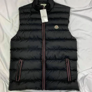 Svart dunväst från Moncler - Snygg svart dunväst från Moncler med quiltad design och ståkrage. Västen har dragkedja framtill med rödvita detaljer och två fickor med matchande dragkedjor. Moncler-logga på bröstet ger en exklusiv touch.