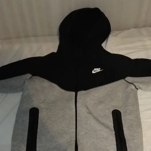 Nike grå och svart hoodiejacka - Nike hoodiejacka i grått och svart med dragkedja framtill och två svarta fickor. Jackan har huva och klassisk Nike-logga på bröstet. Perfekt för en sportig och avslappnad stil.