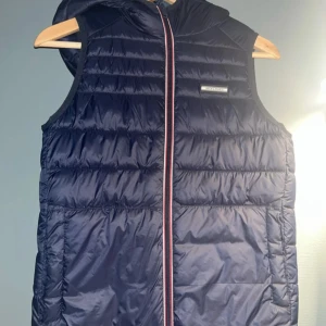 Mörkblå dunväst från Jack & Jones - Snygg mörkblå dunväst från Jack & Jones med huva och cool randig dragkedja i rött, vitt och blått. Västen är quiltad och har en stilren logga på bröstet. Perfekt att slänga över en hoodie för extra värme.