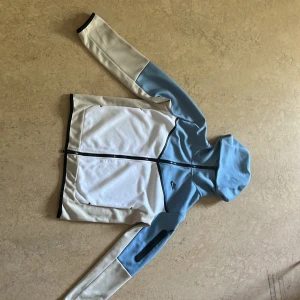 Nike blåvit vindjacka med huva - Nike vindjacka i ljusblått och vitt med svarta detaljer. Jackan har huva, hel dragkedja framtill och en extra ficka med dragkedja på ena ärmen. Priset kan diskuteras vid snabb affär.