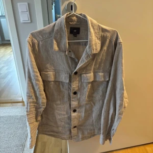 Beige overshirt från H&M i linne - Snygg beige overshirt från H&M i linne med regular fit. Jackan har stora bröstfickor med lock, klassisk krage och knäppning framtill. Perfekt att slänga över en t-shirt för en chill och stilren look. Saknar en knapp på armen