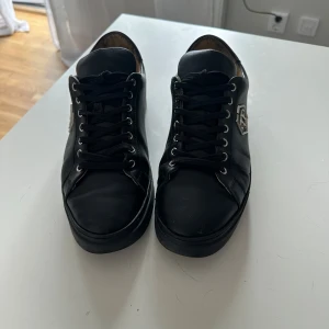 Svarta sneakers från Philipp Plein - Stilrena svarta sneakers från Philipp Plein i skinn med silverfärgad logga på sidan. Klassisk rund tå, svarta snören och metallöglor ger en lyxig känsla. Perfekta för dig som gillar exklusiva detaljer och snygg design.