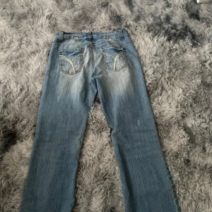 Blå bootcut jeans med broderade fickor - Snygga blå jeans med bootcut passform och coola broderade detaljer på bakfickorna. Jeansen har slitningar och ljus tvätt för en avslappnad vibe. Klassisk femficksmodell med bälteshällor och dragkedja. Perfekta för dig som gillar en retrostil.