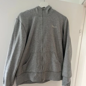 Grå hoodie med dragkedja - Säljer en grå hoodie med hel dragkedja framtill och två stora fickor. Tröjan har huva och är långärmad, perfekt för chill dagar. Enkel och stilren design med liten logga på bröstet. Passar dig som gillar en avslappnad look. Tröjan är i barnstorlek XL men passar xs/s