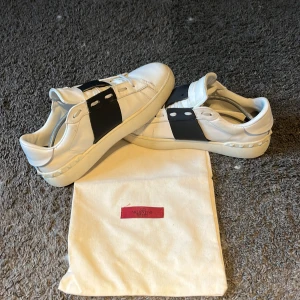 Valentino Open sneakers vit/svart skinn - Inte fast pris!! Hör av er vid minsta fråga Snygga Valentino Open sneakers i vitt skinn med bred svart rem över vristen och ikoniska nitar på hälen. Klassisk rund tå och platt sula. Kommer med originalpåse. Perfekta för dig som gillar lyxiga och stilrena sneakers med en twist. 
