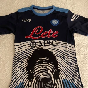 Napoli Maradona 10 fotbollströja EA7 - Napoli fotbollströja med Maradona 10 på ryggen. Mörkblå med ljusblå detaljer och unikt fingeravtrycksmönster framtill. Officiella loggor och sponsorer, tillverkad i 100% mikrofiber. Perfekt för dig som älskar fotboll och Napoli.