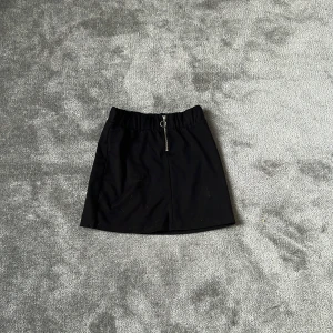 Svart kortkjol från Lindex med dragkedja - Svart kortkjol från Lindex med en snygg silverfärgad dragkedja framtill och elastisk midja. Kjolen är enkel och stilren, perfekt att matcha med en cool t-shirt eller hoodie. Materialet känns mjukt och bekvämt.