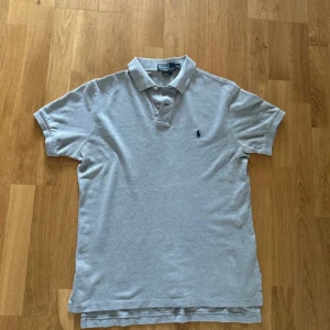 Ralph Lauren - Klassisk grå pikétröja från Polo Ralph Lauren med broderad marinblå logga på bröstet. Tröjan har korta ärmar, krage och knäppning med två knappar. Tillverkad i mjuk bomull och passar perfekt till jeans eller shorts.