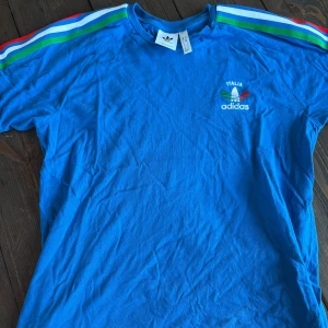 Blå Adidas t-shirt Italia M - Blå t-shirt från Adidas med broderad Italia-logga och färgglada ränder i grönt, blått, vitt och rött längs axlarna. Klassisk passform och rund hals. Perfekt för dig som gillar sportig retrostil.