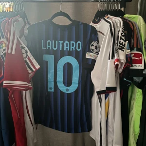 Inter Milan Lautaro Martinez fotbollströja 2025/26 - Inter Milan hemmatröja med Lautaro 10 på ryggen, Champions League-märke på ärmen och sponsortryck från Betsson. Tröjan är svart och blå med smala vertikala ränder, klassisk rund hals och tillverkad i lätt funktionsmaterial. Perfekt för dig som älskar fotboll och vill sticka ut på ett positivt sätt. 