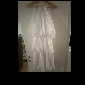 Vit kort klänning med volanger - Superfin vit kort klänning med flera lager volanger och glittrigt tyg. Klänningen har halterneck design med snygga guldknappar och plisserad struktur. Perfekt för dig som vill sticka ut med en romantisk och flowy look.